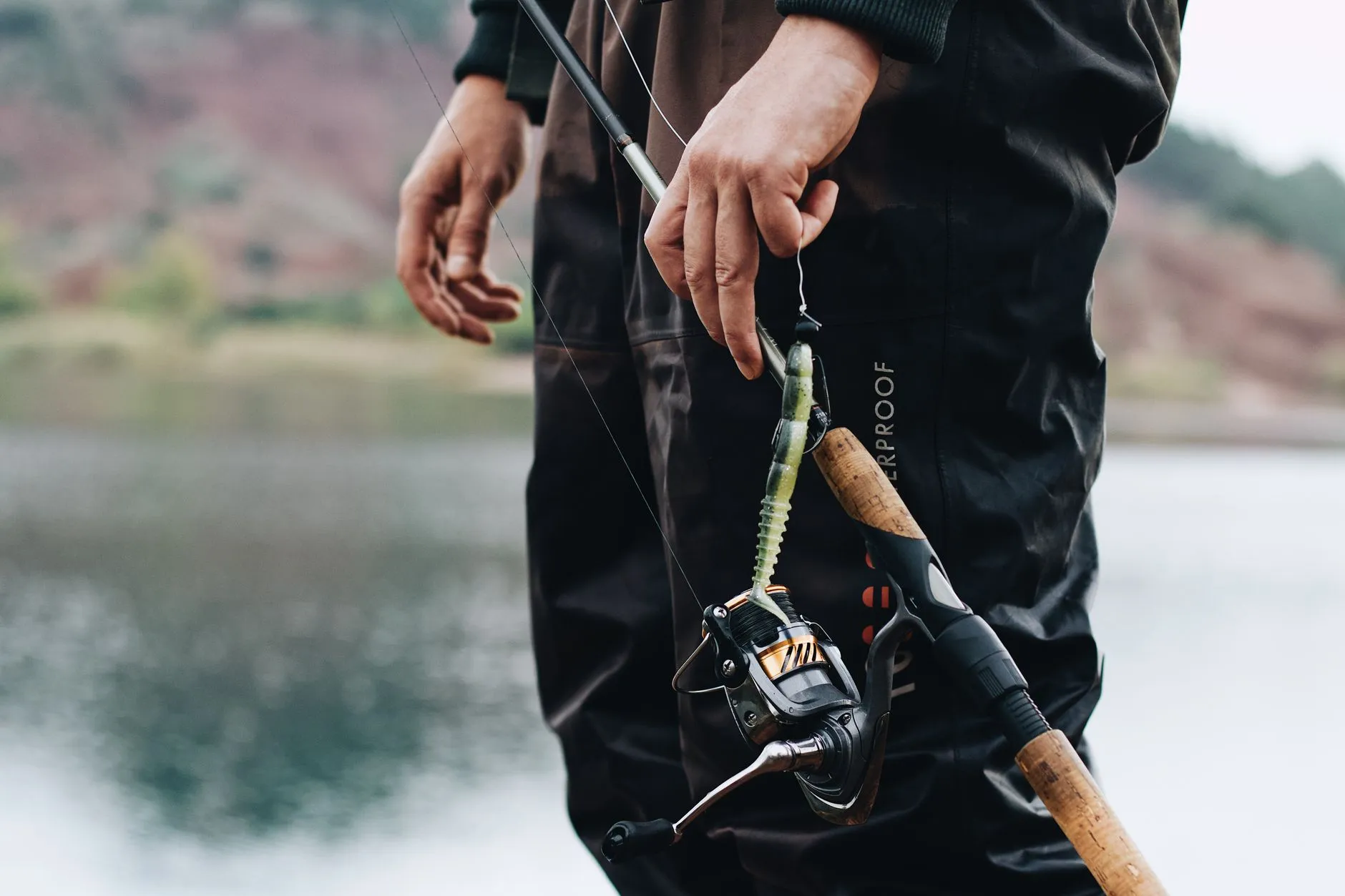 Angler in wasserdichter Wathose hält eine Rute mit Stationärrolle am Ufer eines Sees
