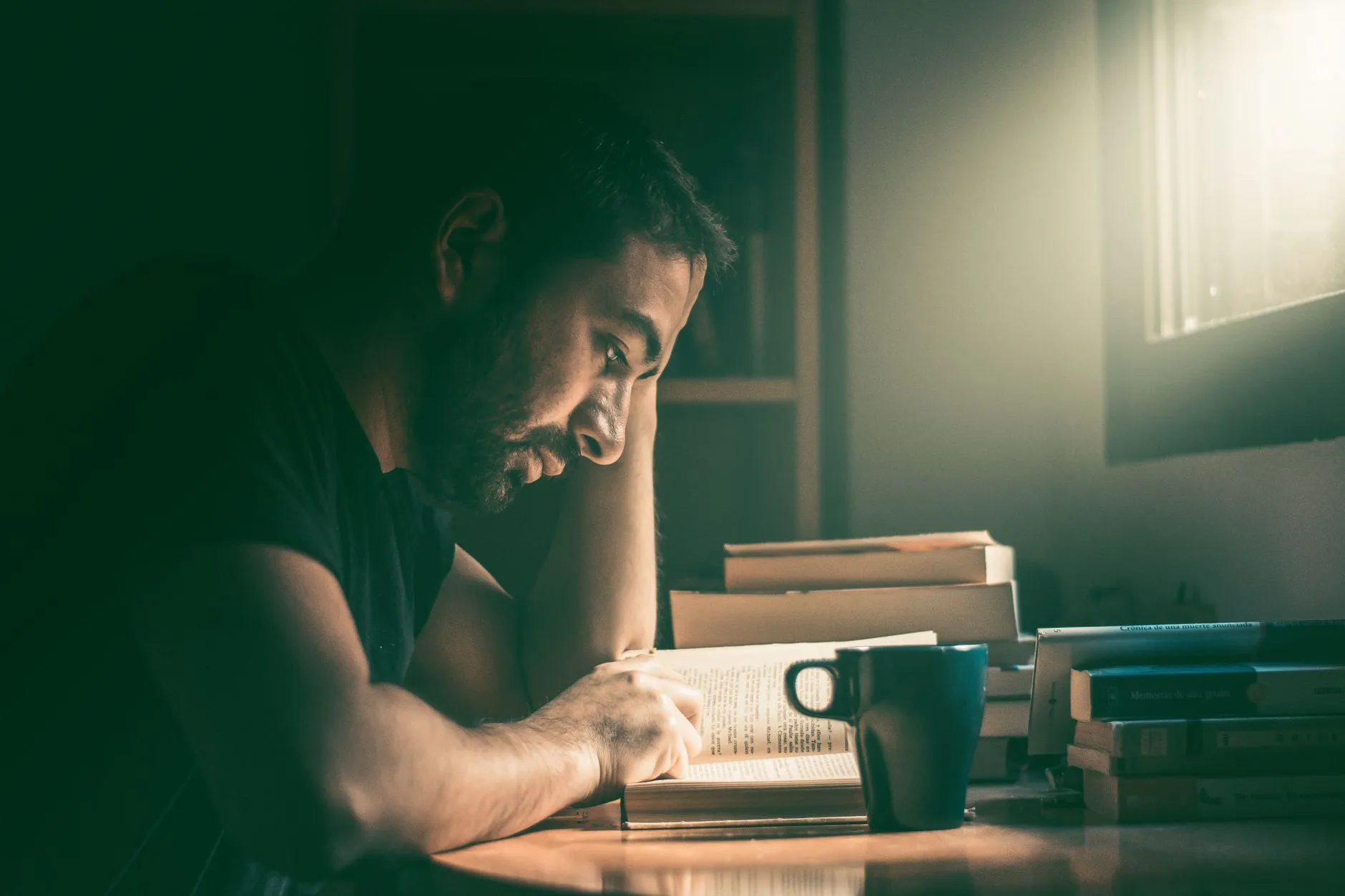 Mann sitzt am Tisch und liest in einem aufgeschlagenen Buch, daneben Bücherstapel und eine Tasse