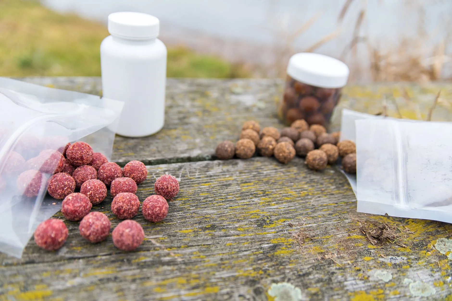 Rote und braune Boilies auf einem verwitterten Holztisch, daneben Plastikbeutel und zwei Kunststoffdosen