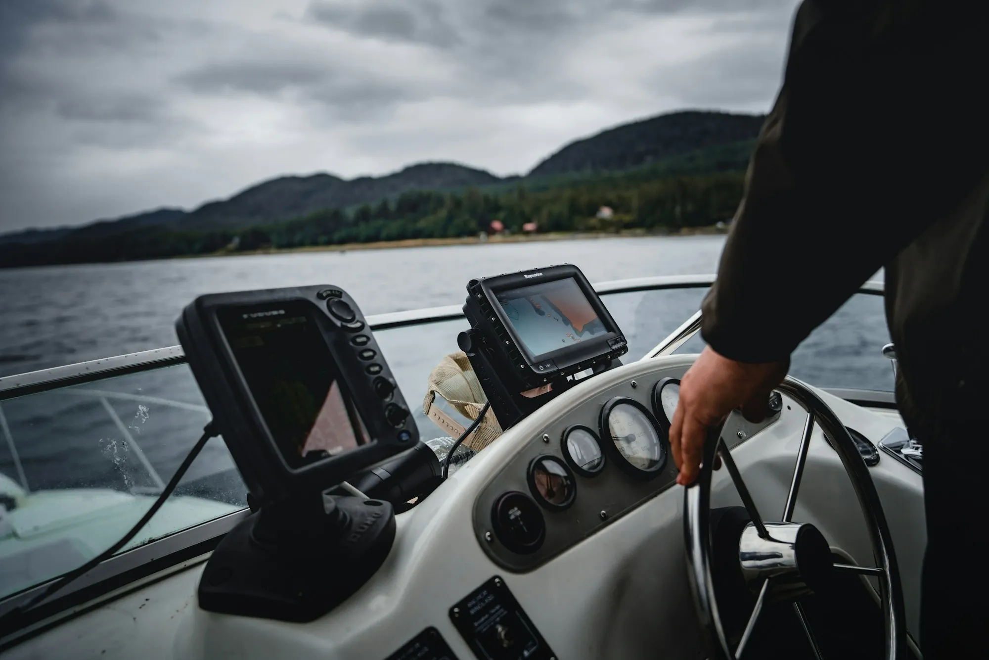 Zwei Bildschirme für Echolot und Navigation am Steuerstand eines Boots, eine Hand liegt am Steuerrad, dahinter eine bewaldete Küste