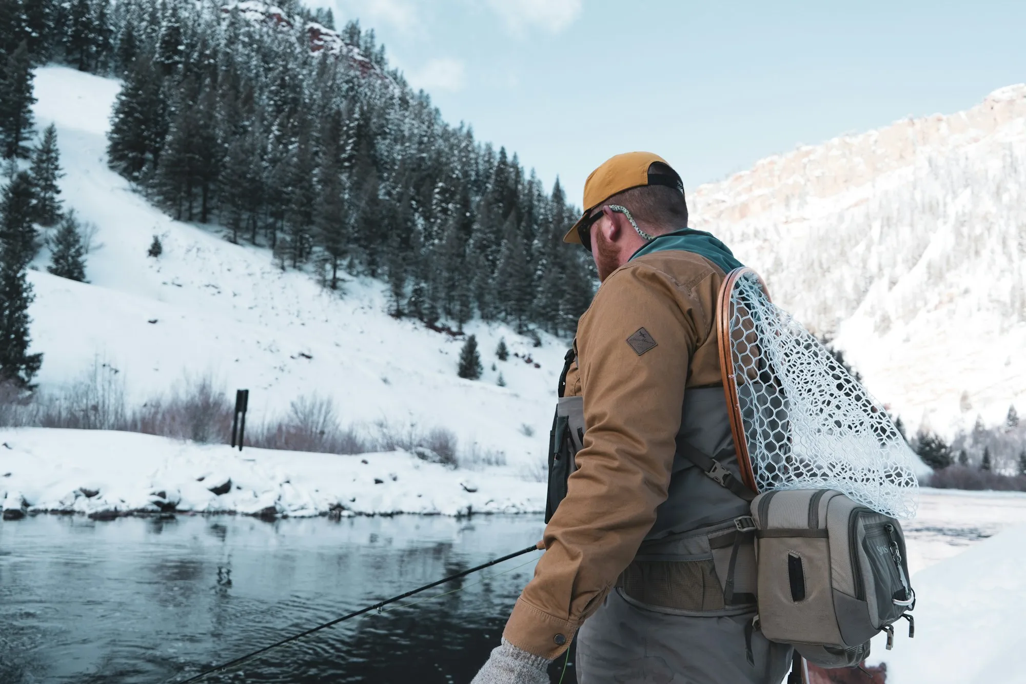 Angler mit Kescher auf dem Rücken steht an einem Fluss in verschneiter Berglandschaft